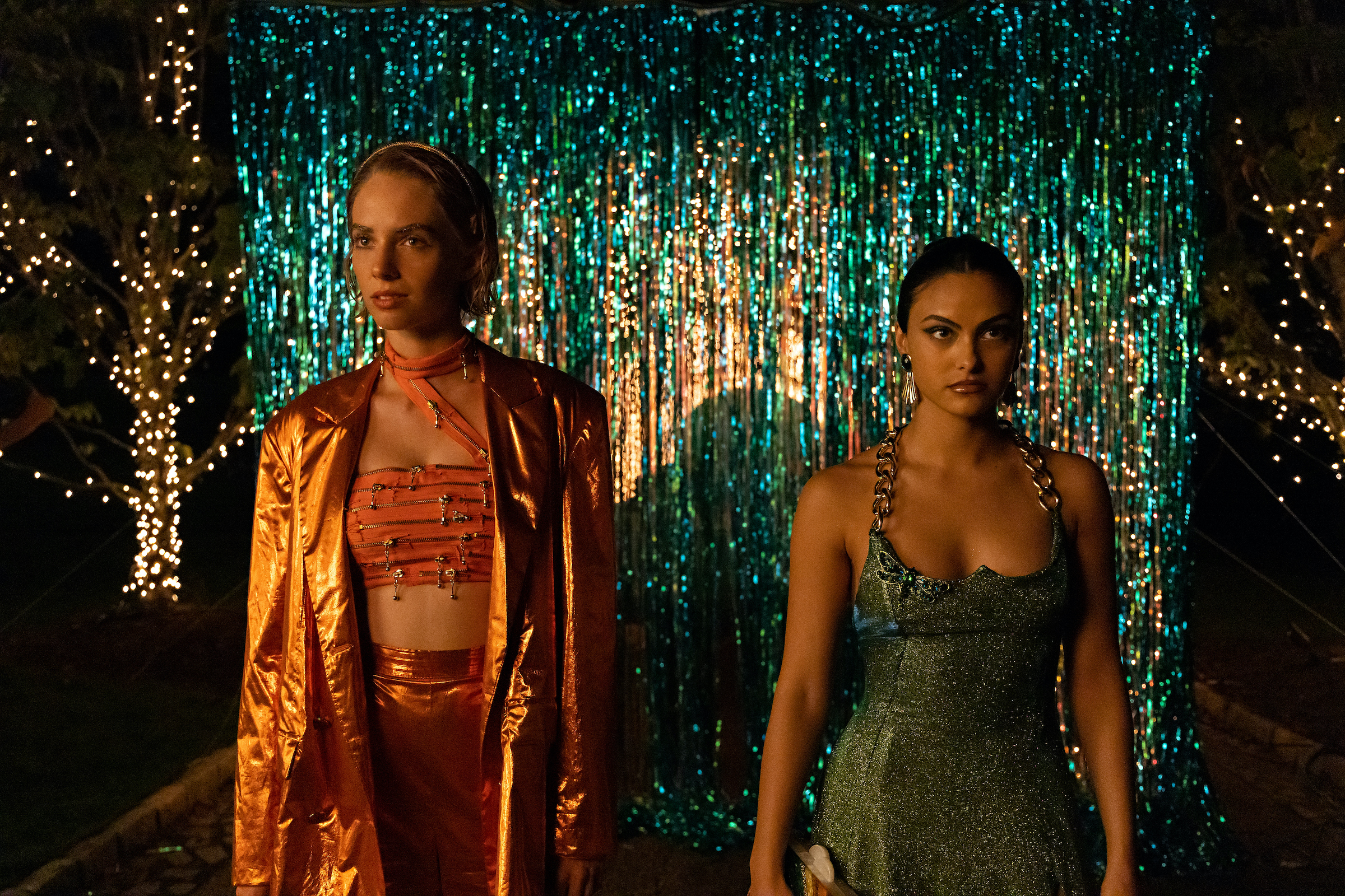 Maya Hawke as Eleanor wears an orange crop top full of zippers under a foil orange suit and Camila Mendes as Drea wears a blue sequined dress with a chain halter neck.