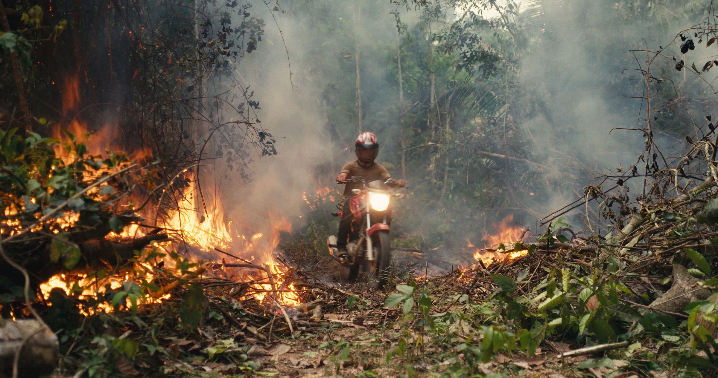 A man on a red motorcycle rides toward the camera through fire on both sides