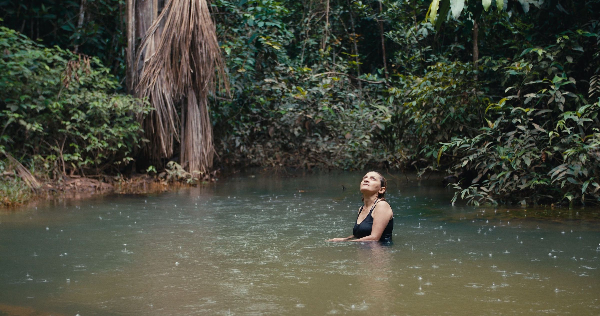 A woman in a black one piece swimsuit stands up to her waist in the river, looking upward