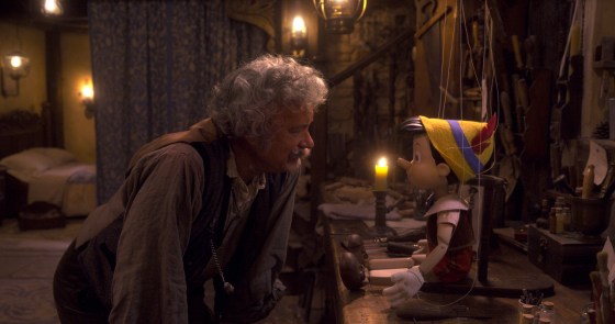 Tom Hanks in a curly gray wig rests his hands on his knees as he squats down to talk to Pinocchio.