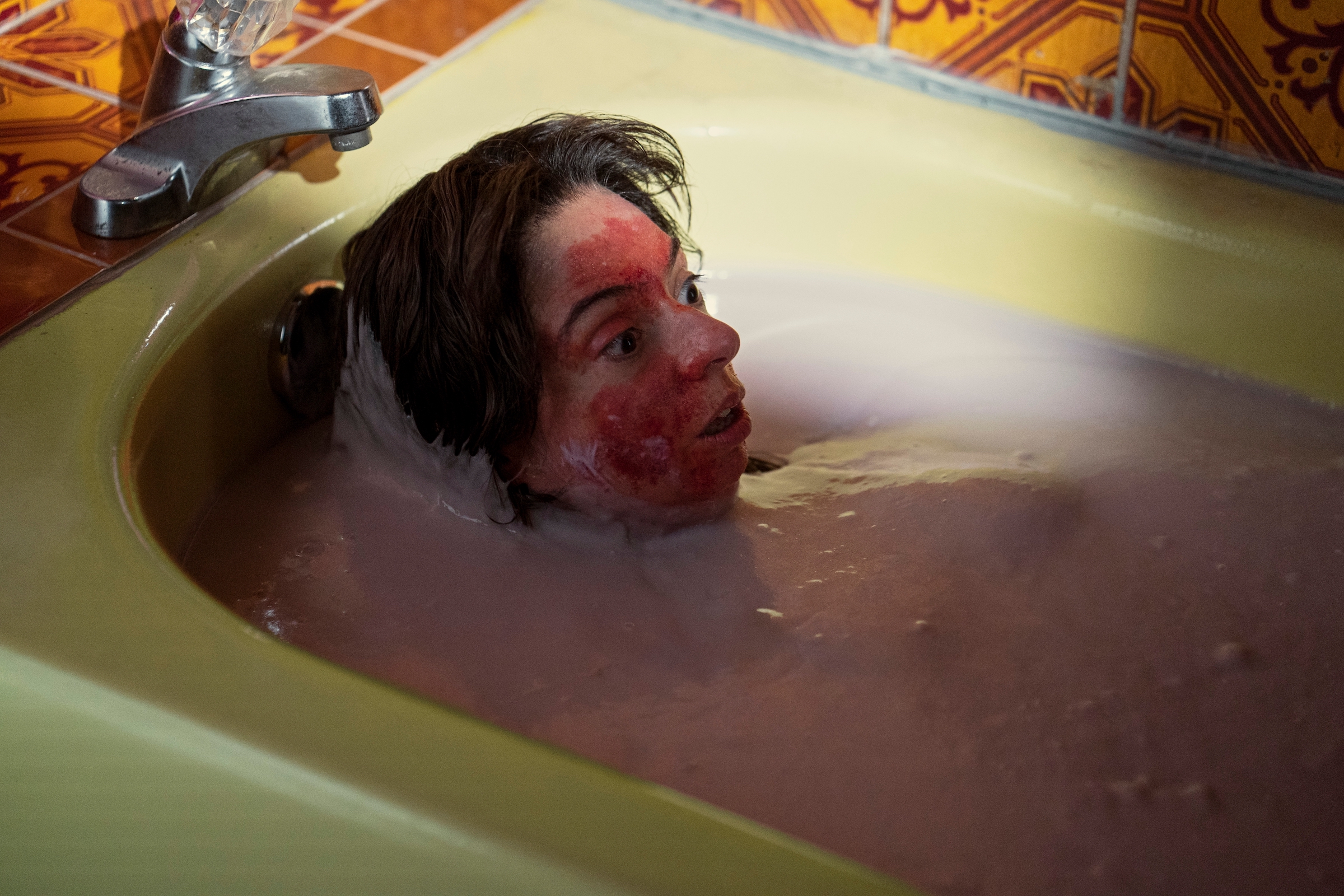 A woman with red smeared all across her face submerges herself in a thick pink bath, looking shocked