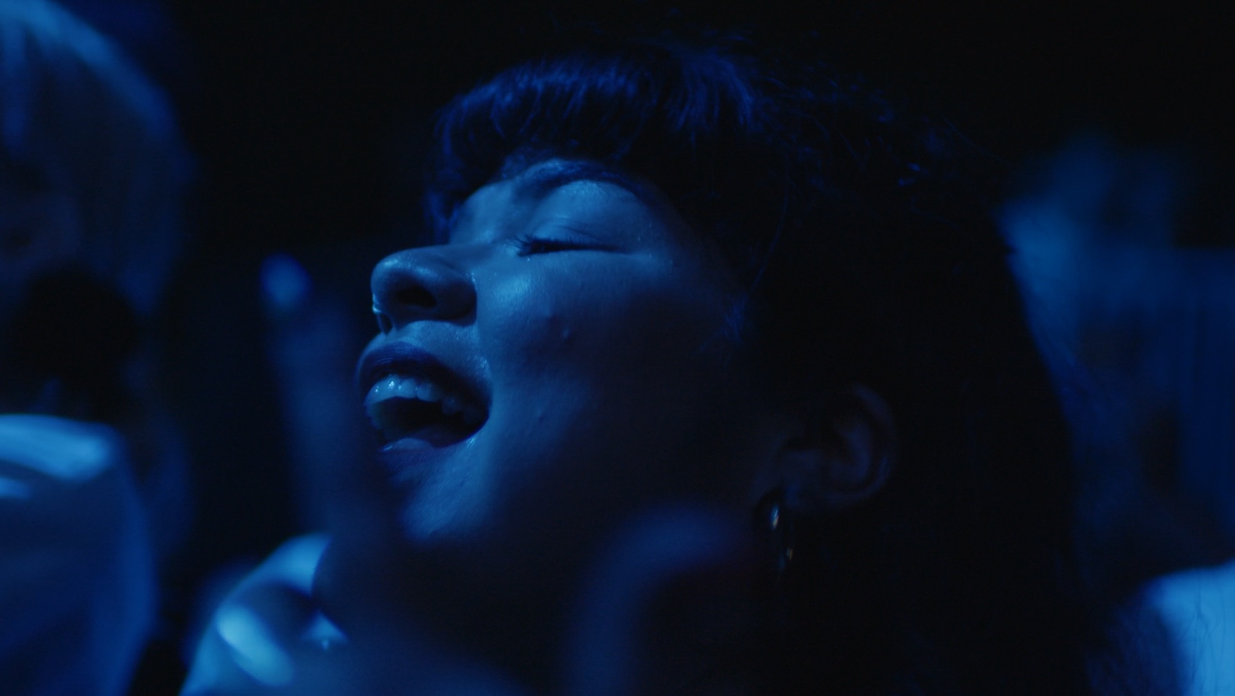 Doris Muñoz dances in a blue-lit crowd at a concert in New York City. This is a close-up of her face grinning, open mouthed.