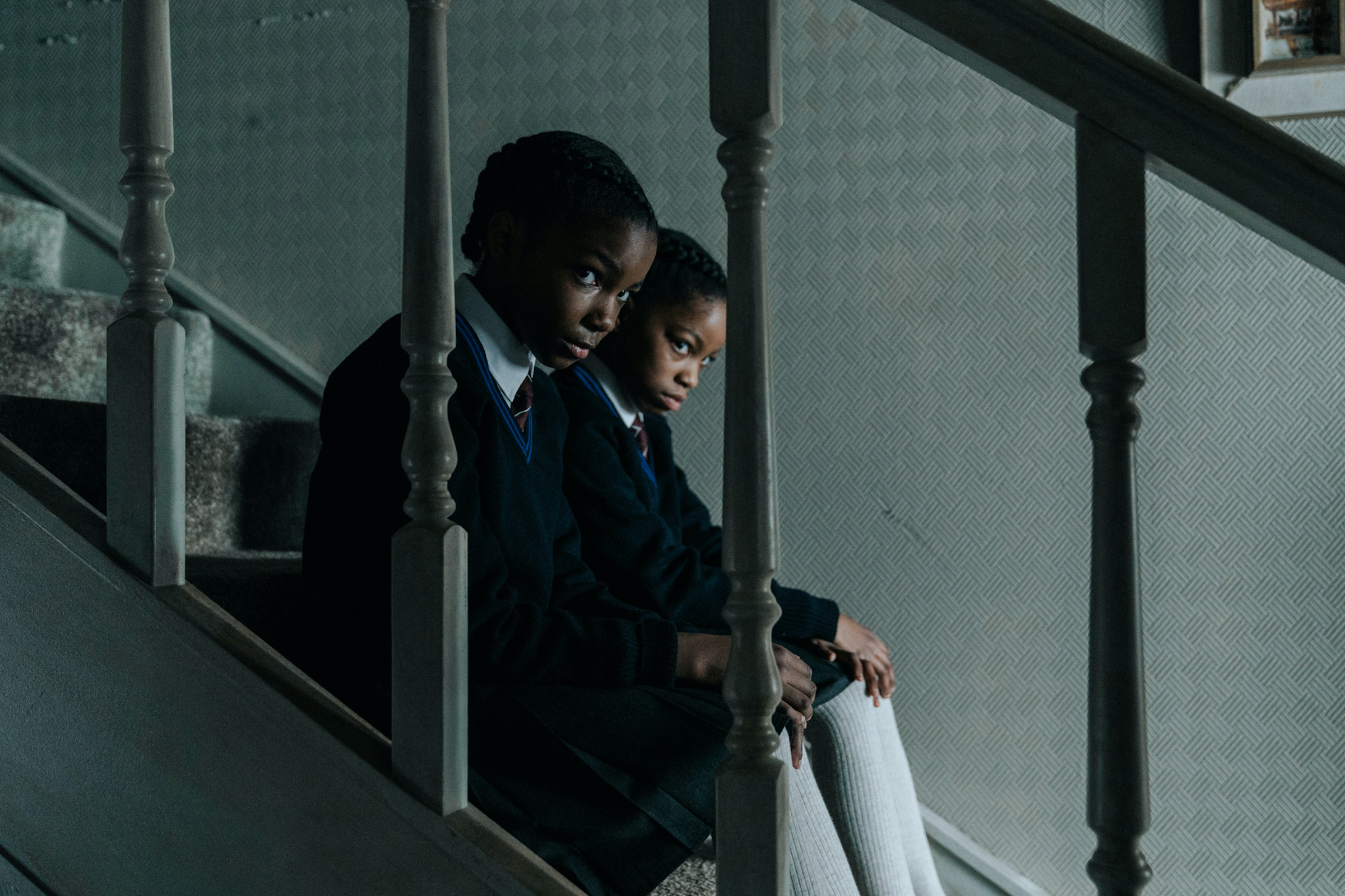 Leah Mondesir Simmons as young June Gibbons and Eva-Arianna Baxter as young Jennifer Gibbons sit on the stairs, peering out through the bannisters.