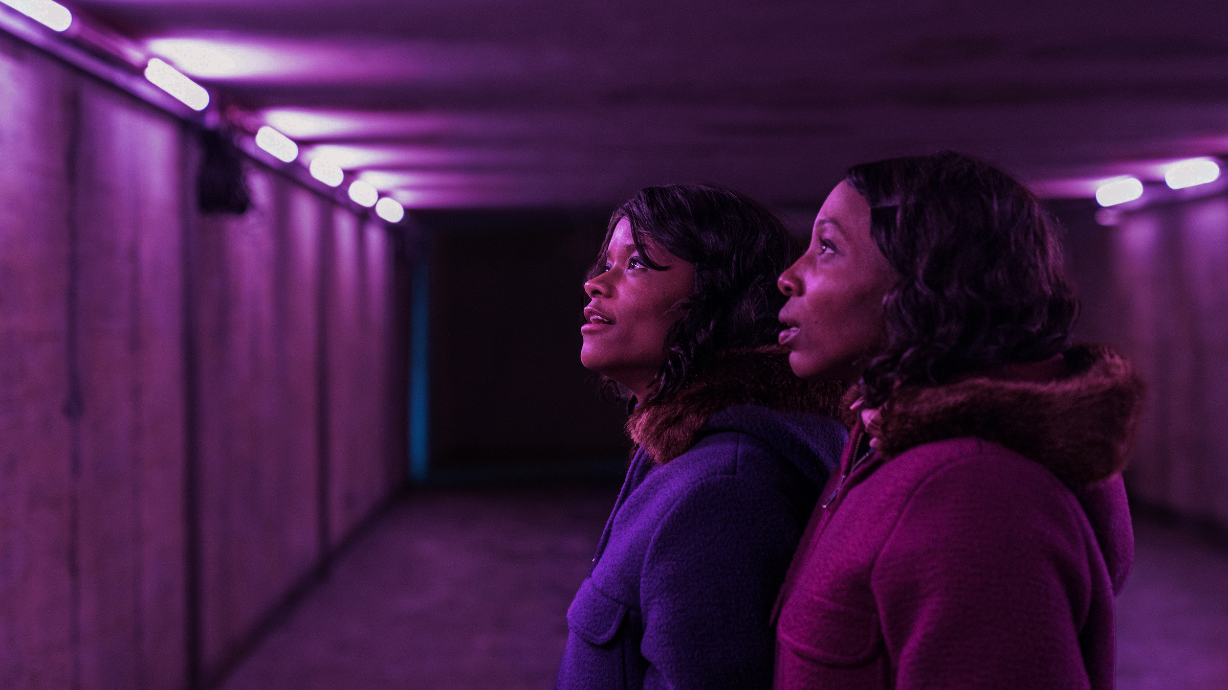 Letitia Wright as June Gibbons and Tamara Lawrance as Jennifer Gibbons gaze up at the purple lights in a concrete underpass