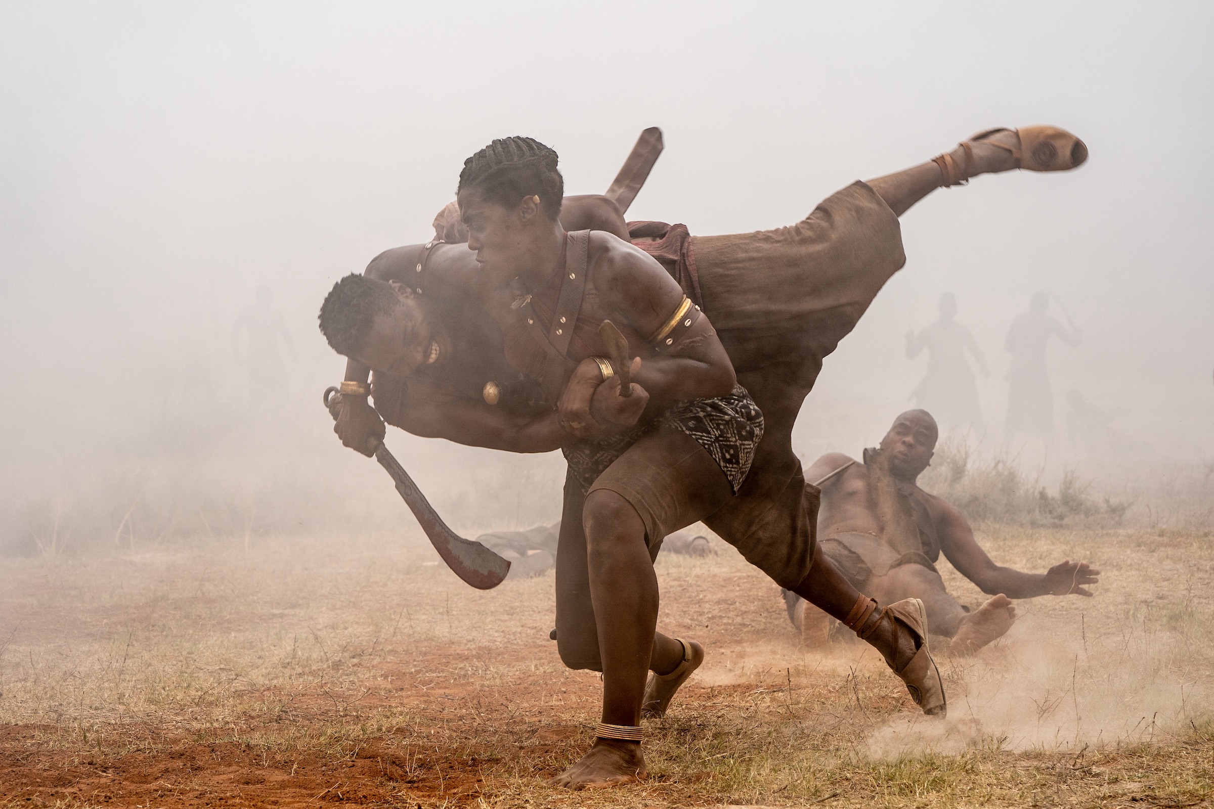 An African woman warrior tackles an African man onto the ground, sword wielded, in battle.