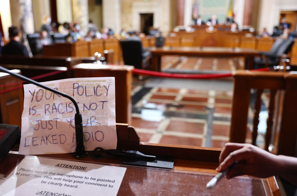 A sign rests against the microphone stand that says “Your Policy Is Racist Not Just Your Leaked Audio,” as protestors demonstrate at the L.A. City Council.