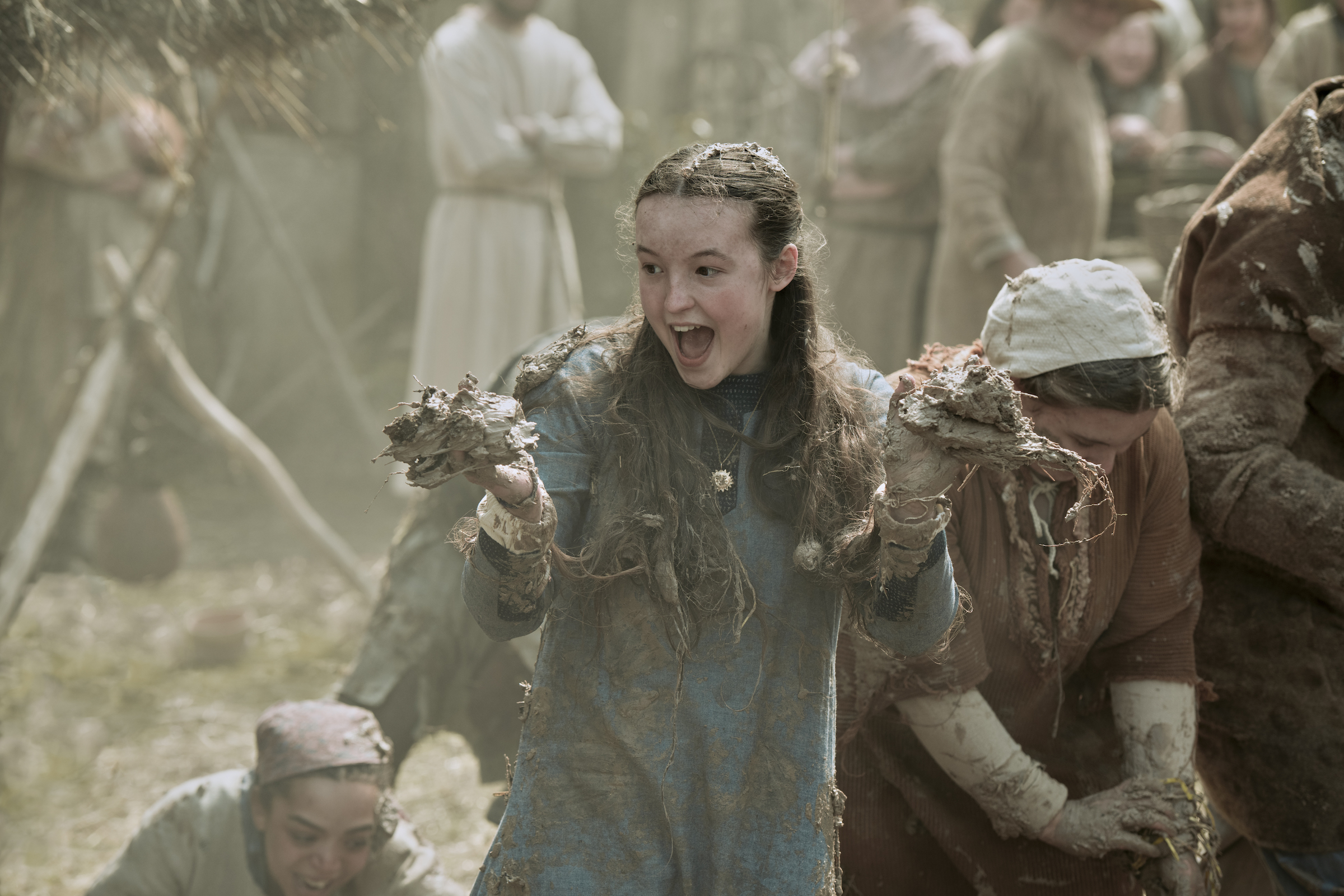 Birdy, in a longsleeve blue dress, grabs fistfuls of mud at a village cottage raising