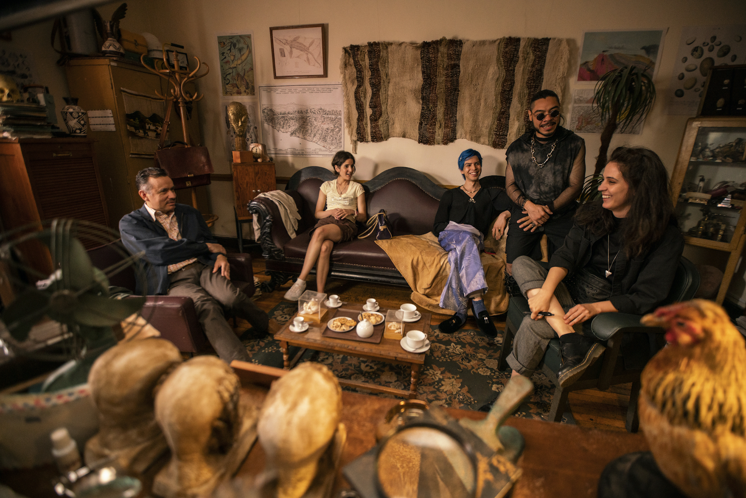 The Espookys and Tío Tico sit in a professor's study around a coffee table
