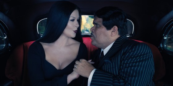Morticia and Gomez clasp hands and lock eyes in the backseat. They look like they're about to kiss.