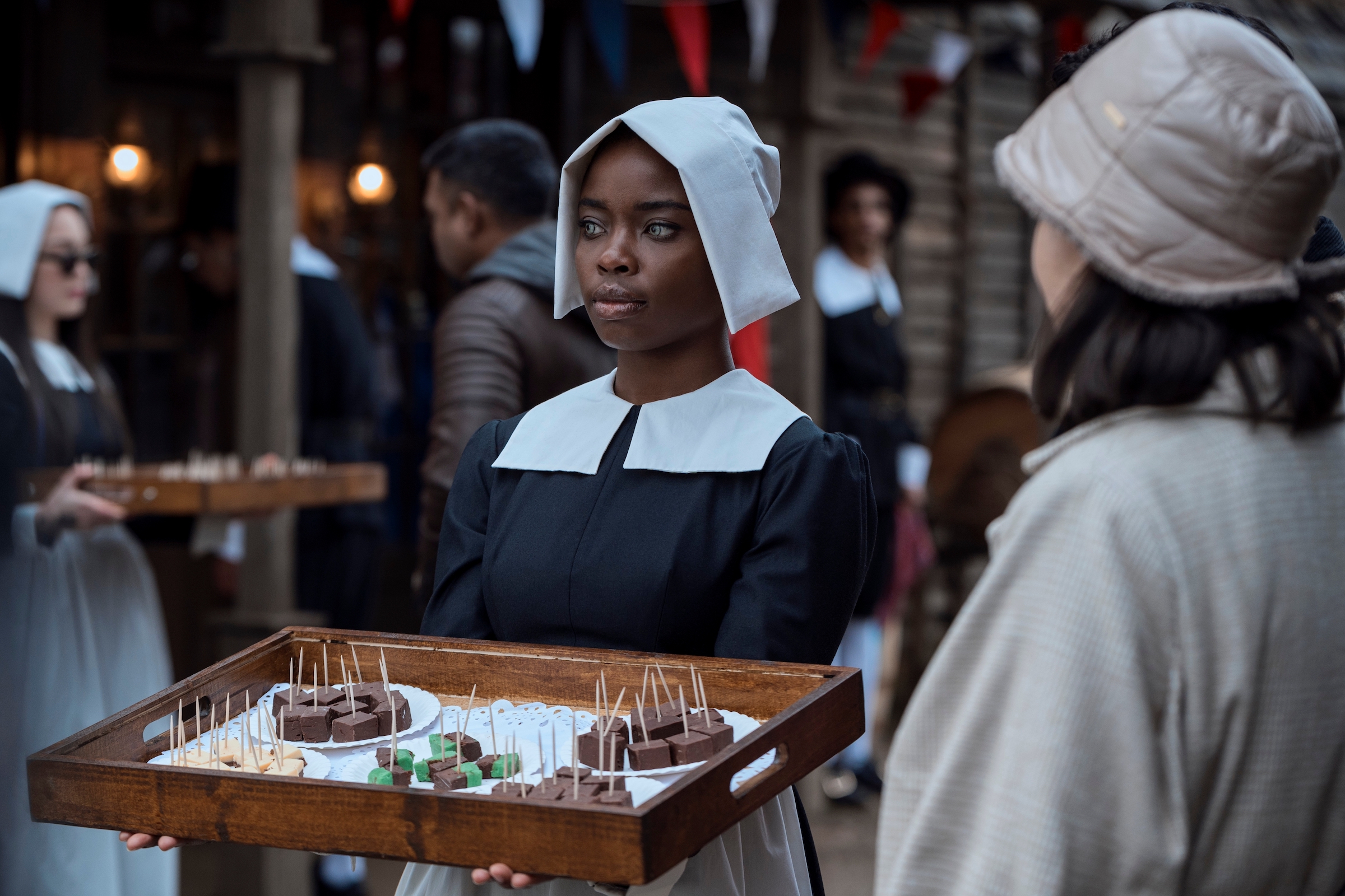 Bianca Barclay (Joy Sunday), wearing a black and white pilgrim outfit, holds out a wooden tray of fudge samples with toothpicks in them.