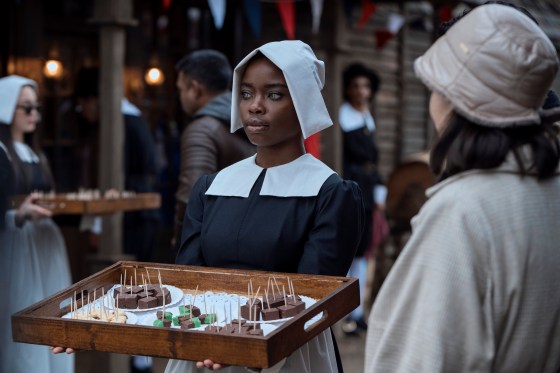 Bianca Barclay (Joy Sunday), wearing a black and white pilgrim outfit, holds out a wooden tray of fudge samples with toothpicks in them.