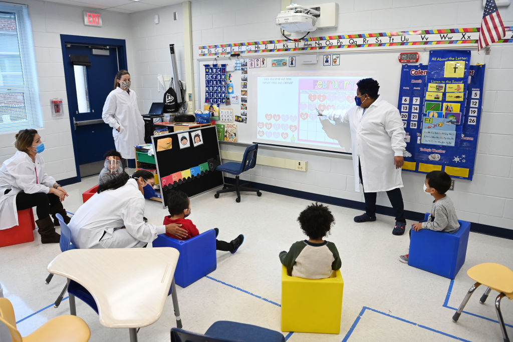 A teacher dressed up in a white lab coat and a mask teaches her preschool autism class with four students sitting in a semicircle around awhiteboard. There are three other teachers also dressed in white lab coats and masks—two are listening to the class and one is talking to one of the children with her hand placed on his left shoulder.