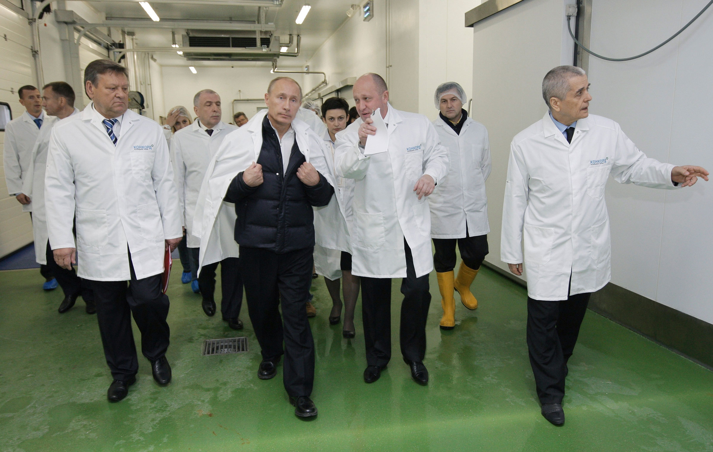 Yevgeny Prigozhin and Vladimir Putin wearing white lab coats and standing with a group of men as they tour a factory in 2010
