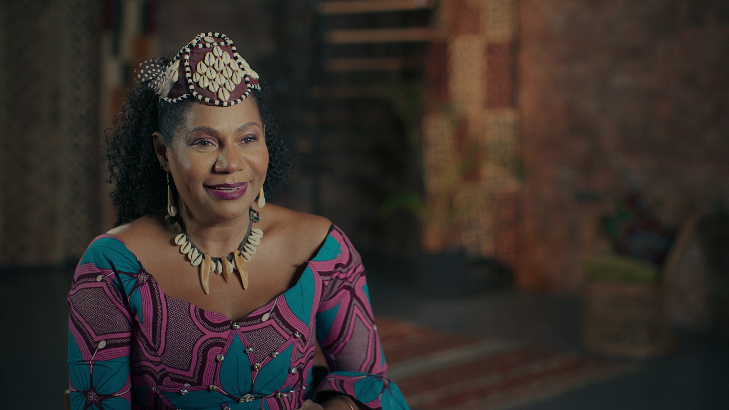 Queen Kabatusuila wears a brown headpiece covered in cowry shells.