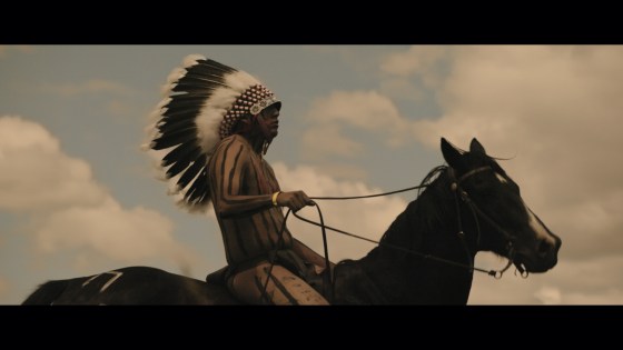 A Native American man in a feathered headdress and striped body paint sits atop a horse, holding the reins up.