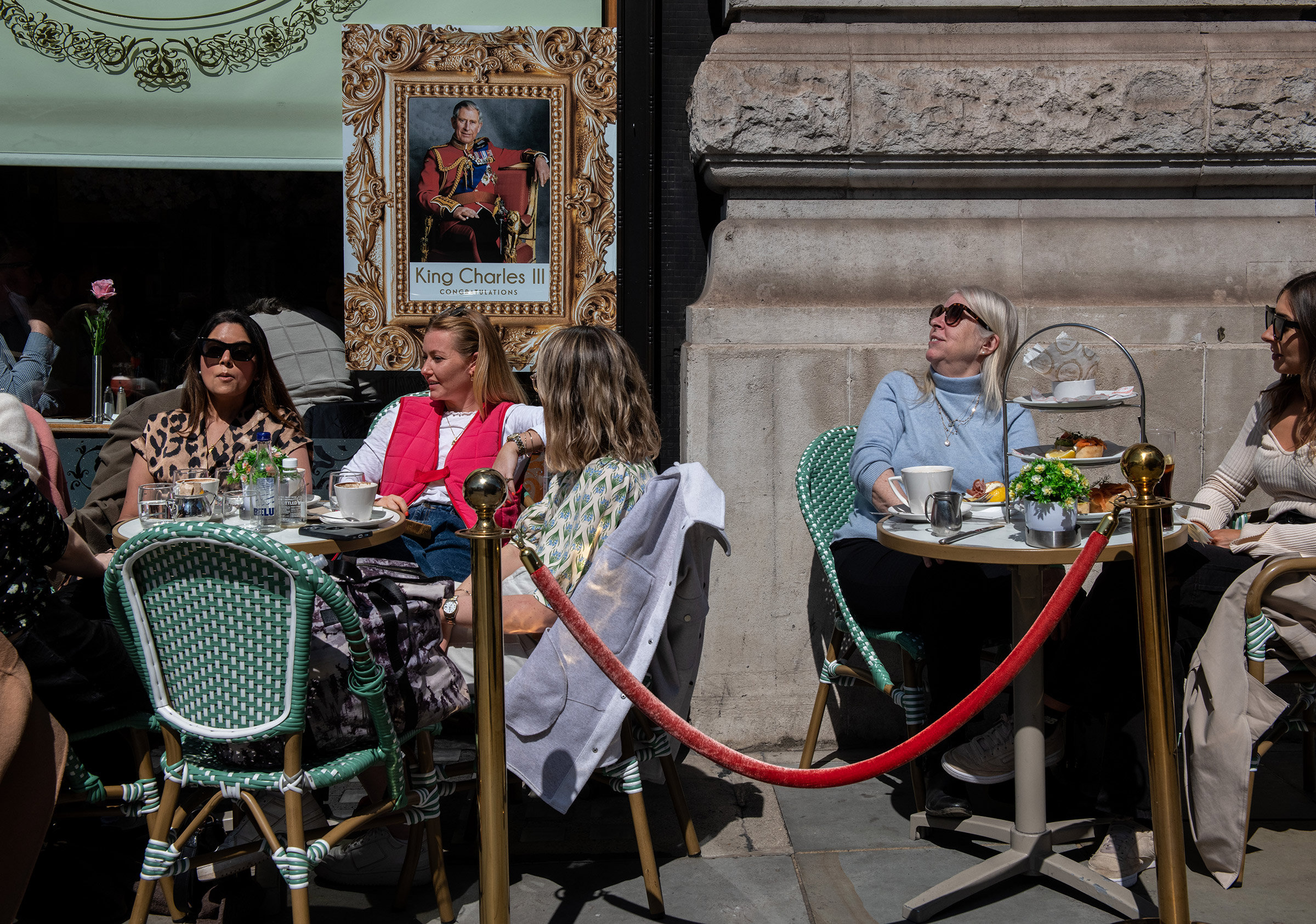 People have afternoon tea outside a cafe next to a poster of King Charles III on April 29.