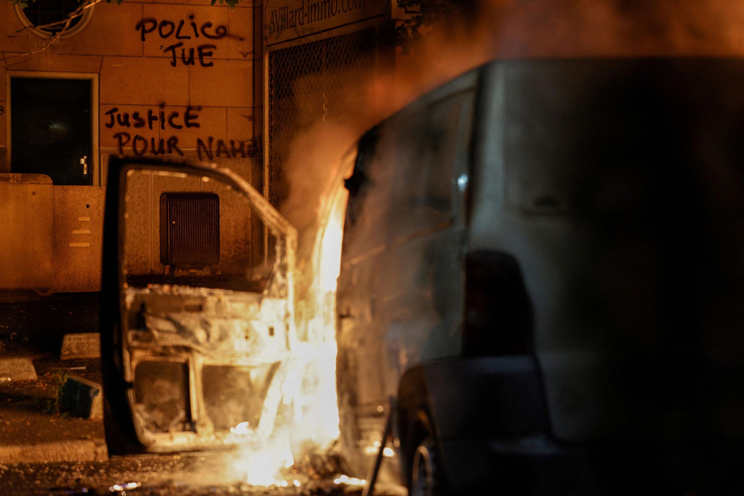 A car burns as slogans are seen on a wall which reads in French “Police kills, Justice for Nahel” during protests in Nanterre