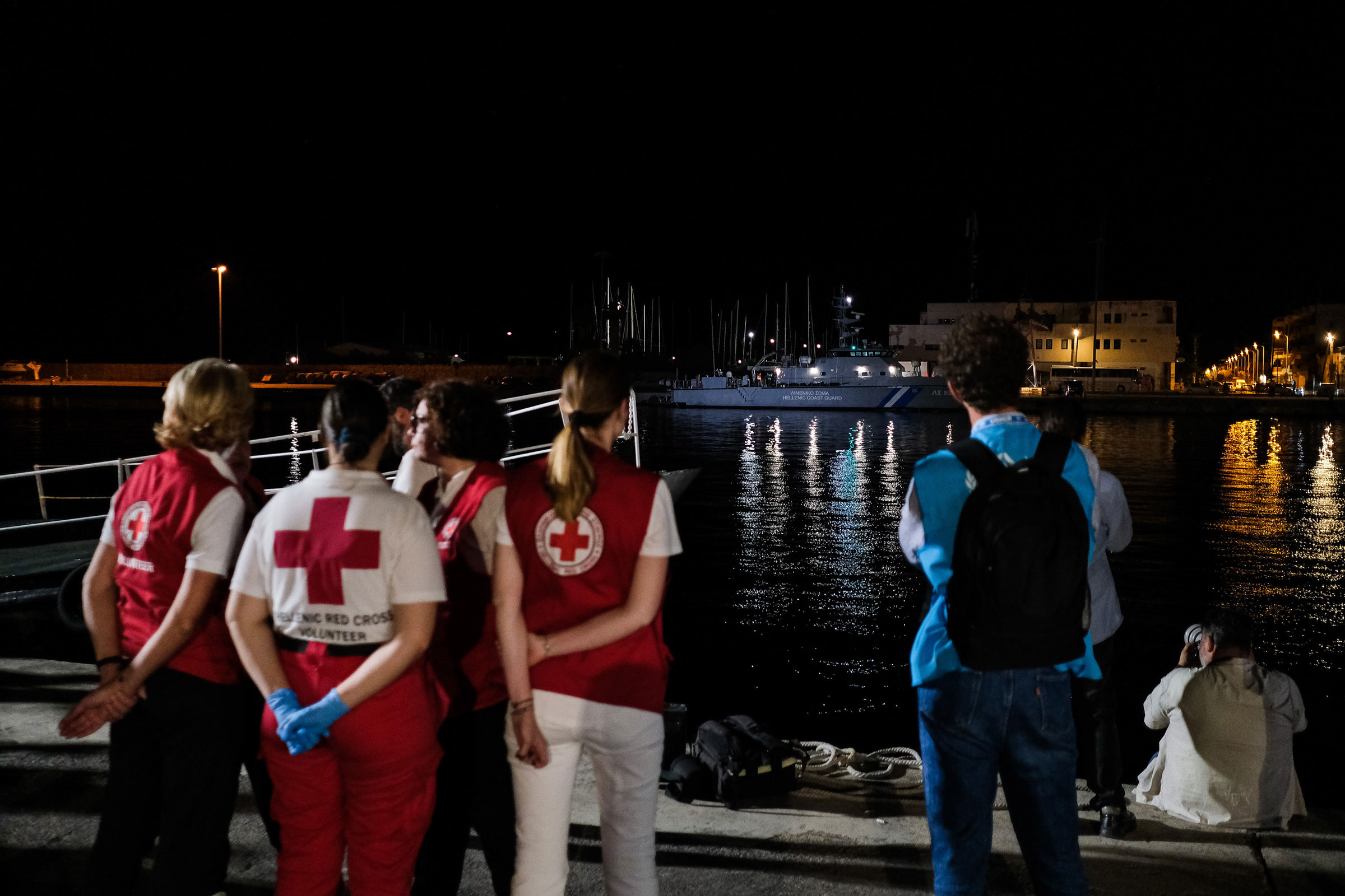 A group of people wearing Red Cross and UNHCR vests stand on a port facing the ocean with their arms crossed as a large boat arrives that is holding the 79 recovered bodies from the wreck