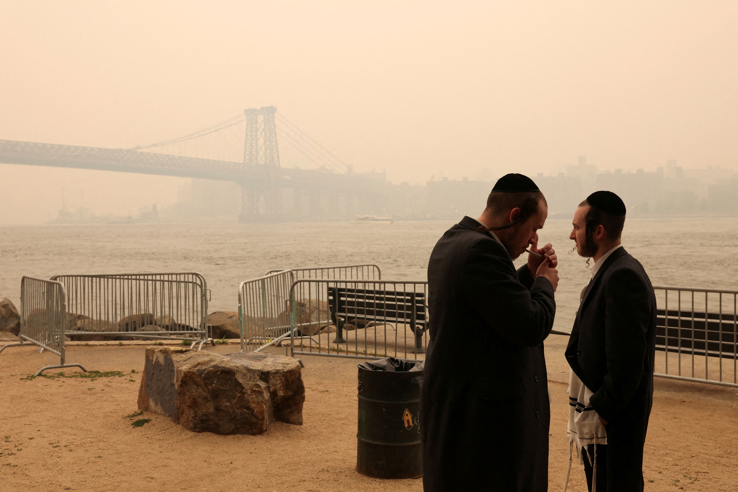 An Orthodox Jewish man lights a cigarette while standing with another by the waterfront as haze and smoke caused by wildfires in Canada cover the Manhattan skyline on June 7, 2023. (Amr Alfiky—Reuters)