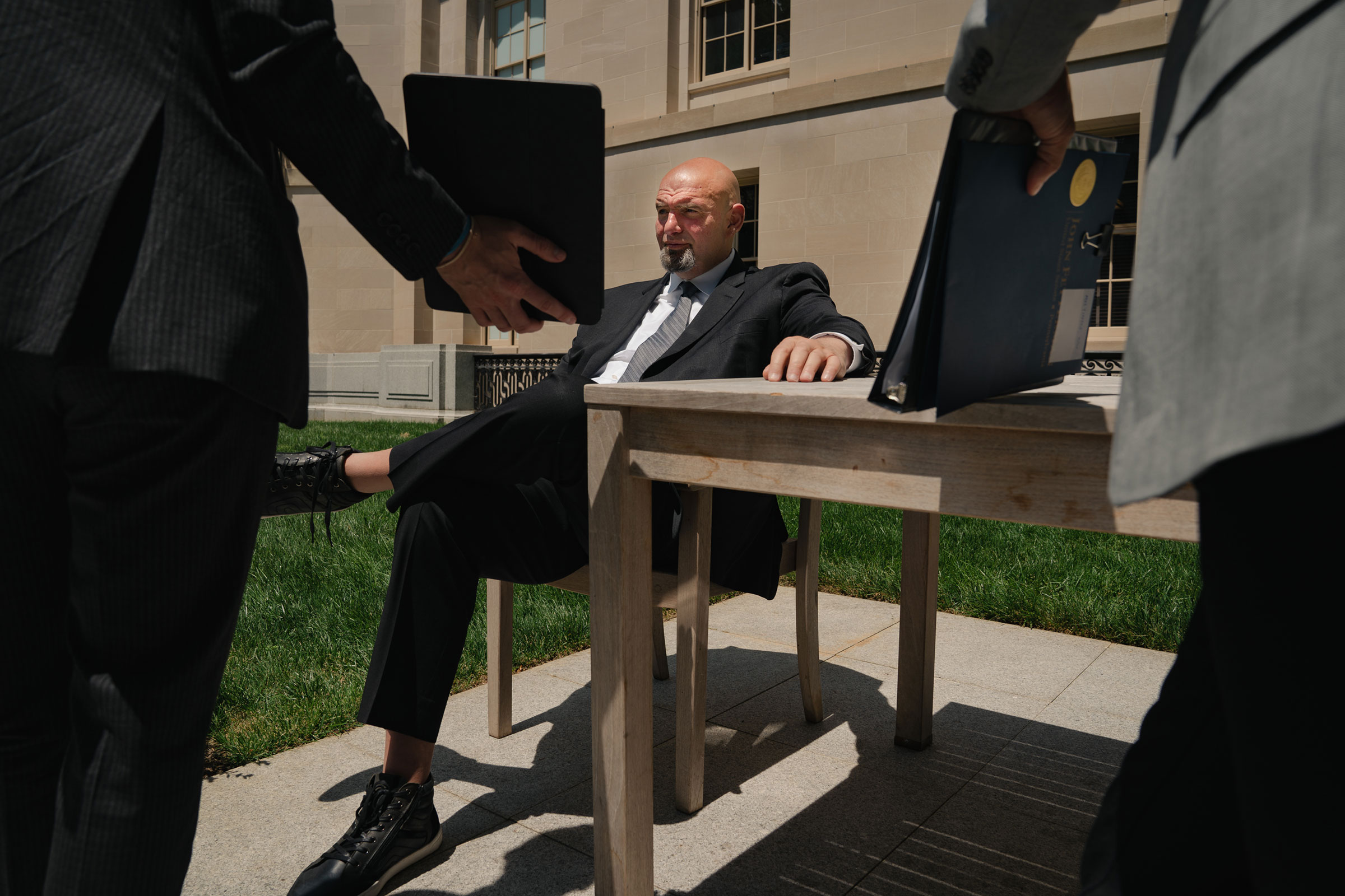 Senator John Fetterman sits in a chair outside in a courtyard in the sun as two other men show him a binder and an ipad, which has a prompter on it so he can have captions of what people around him are saying