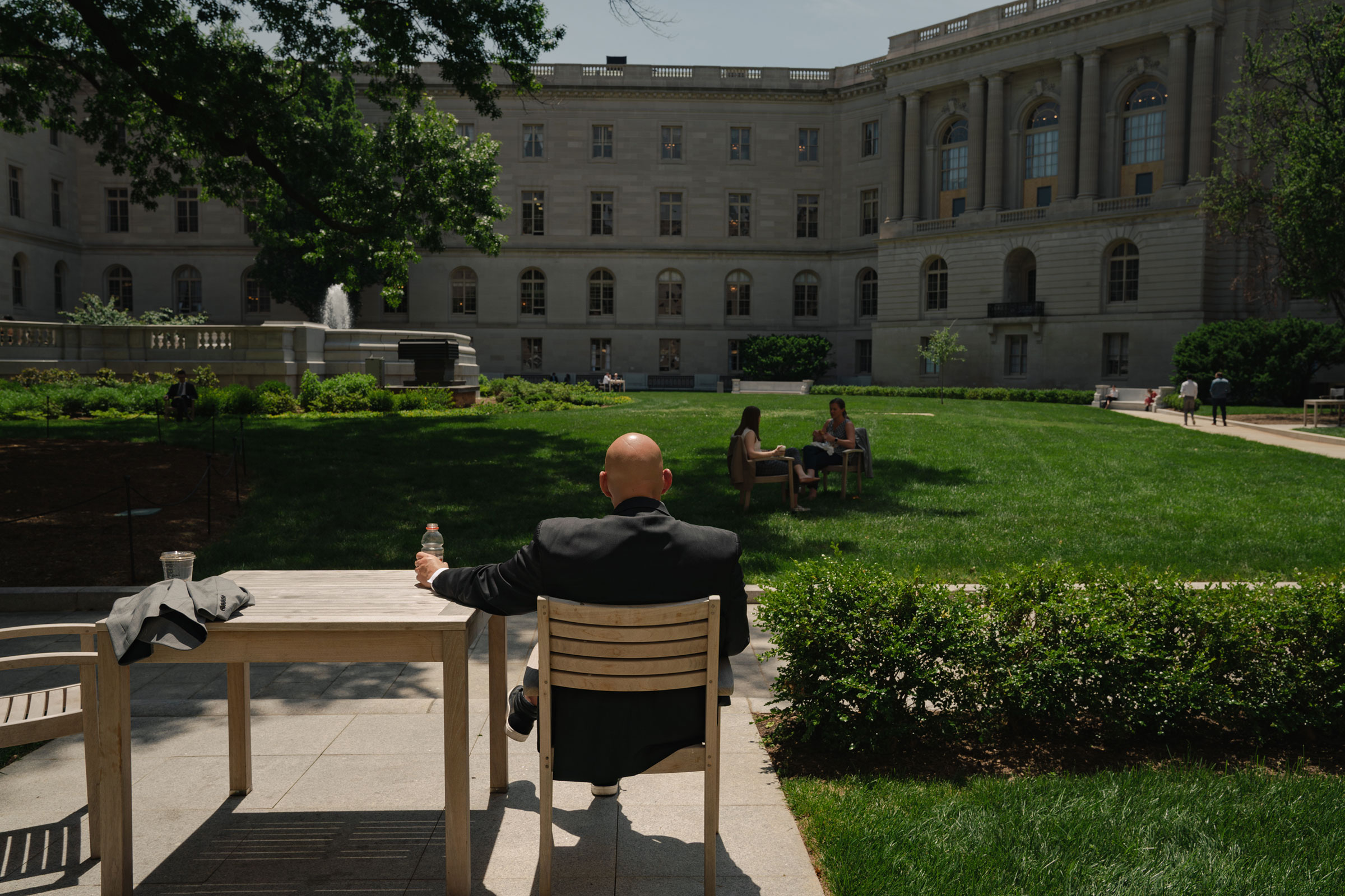 Senator John Fetterman sits in a chair outside in a courtyard at capitol hill with his back to the camera, he is holding a bottle of water resting his arm on a table and looking out into the courtyard