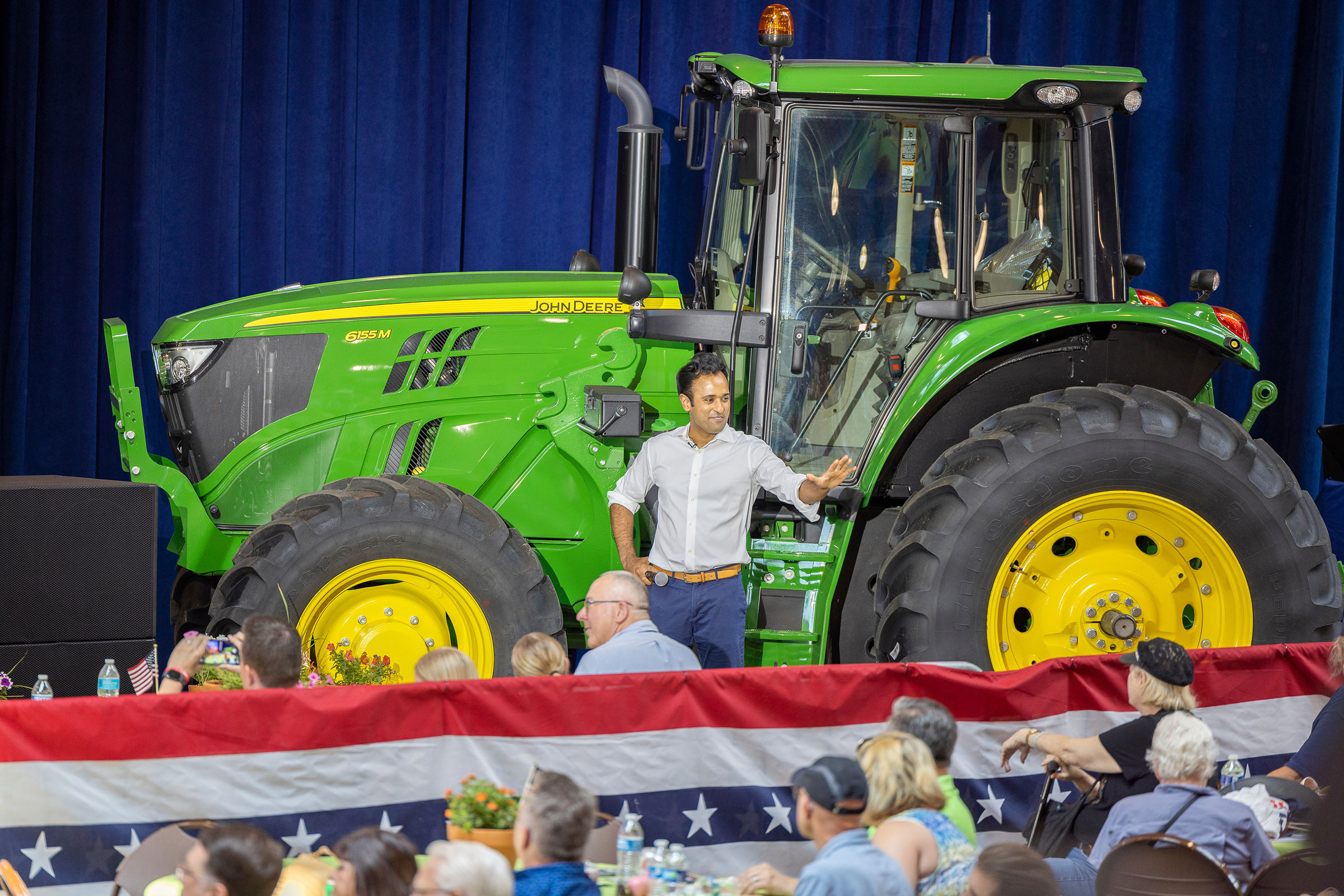 Ramaswamy stands in front of a large green tractor as he waves to supporters in a crowd in front of him