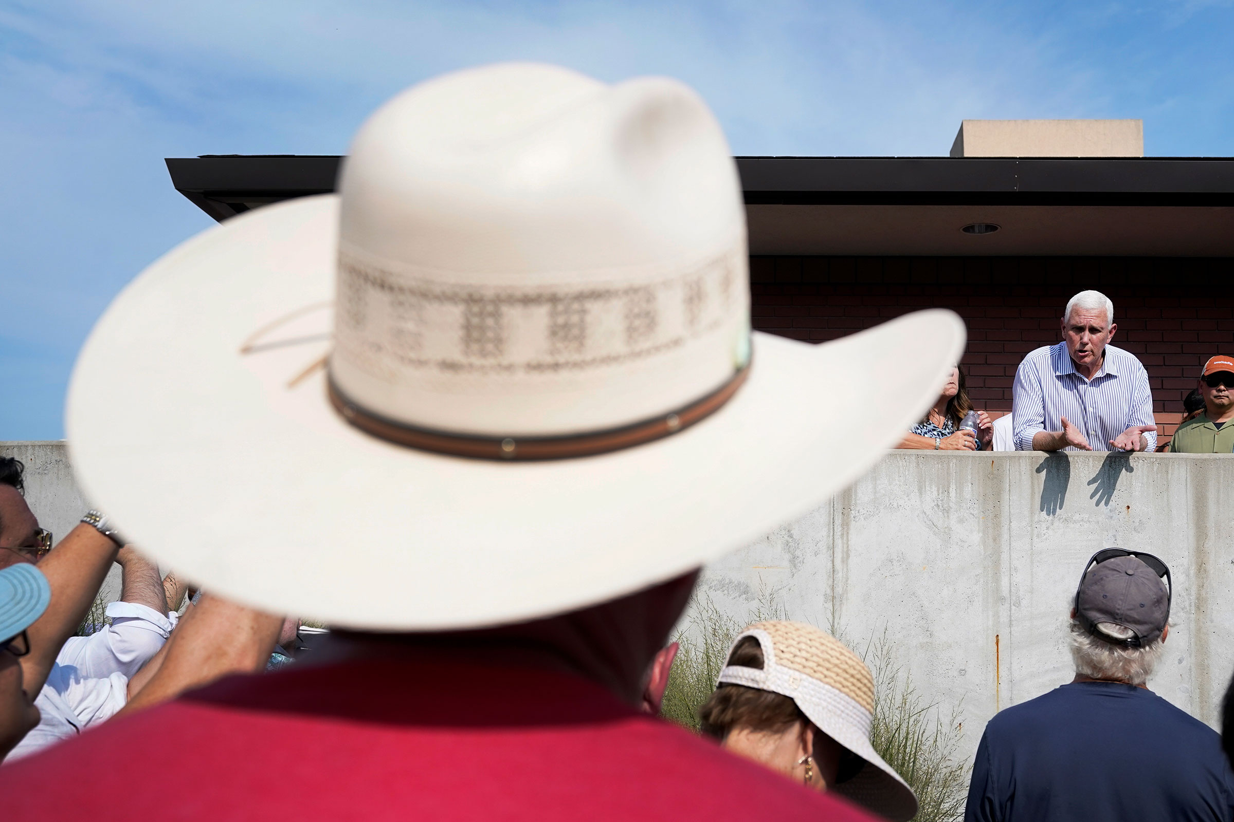 In the foreground a man in a cowboy hat listens to former Vice President Mike Pence as he leans over a concrete wall as he speaks
