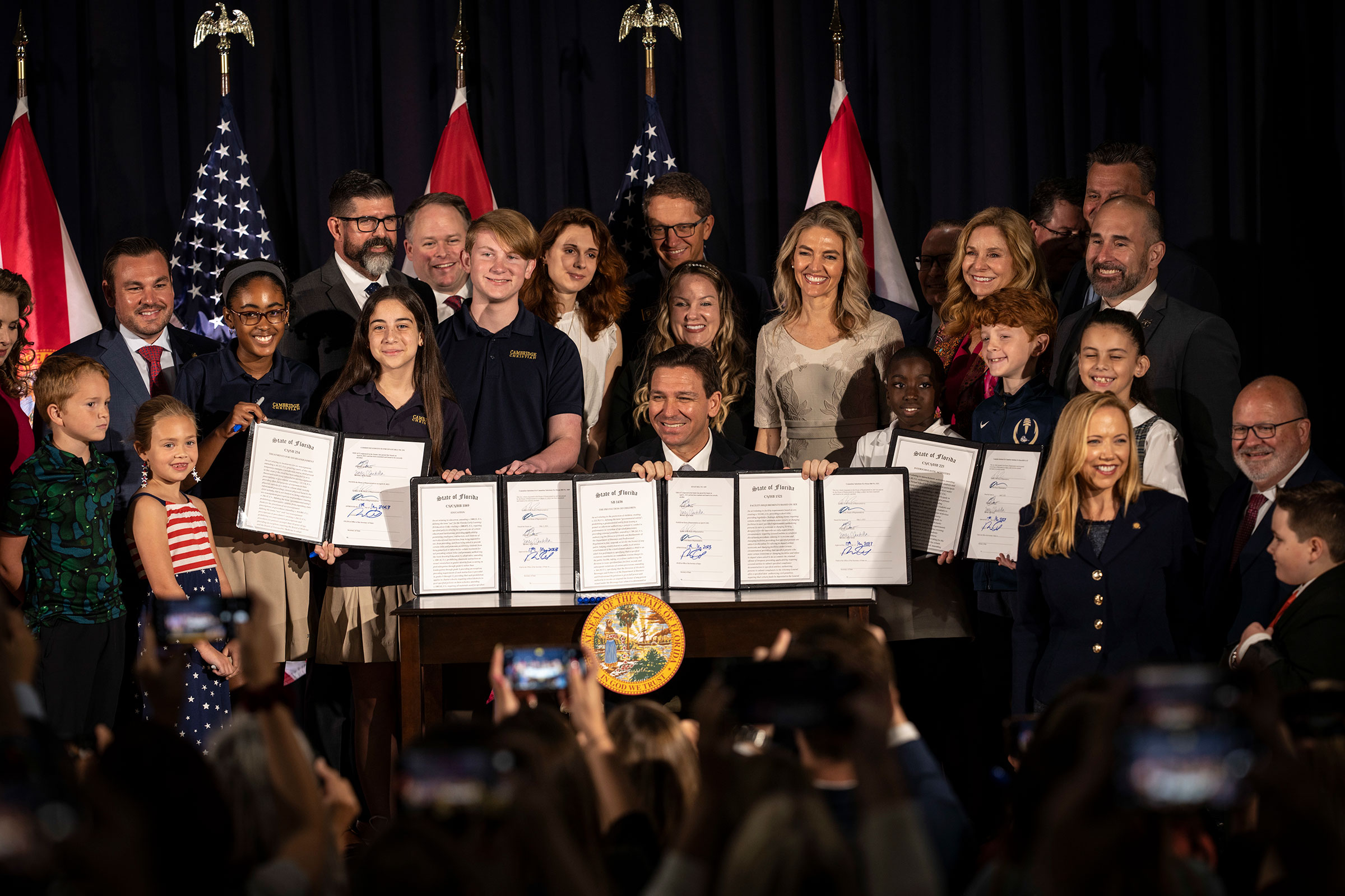 Ron Desantis sits at a table with a group of adults and children standing behind him as he holds up signed bills for the camera