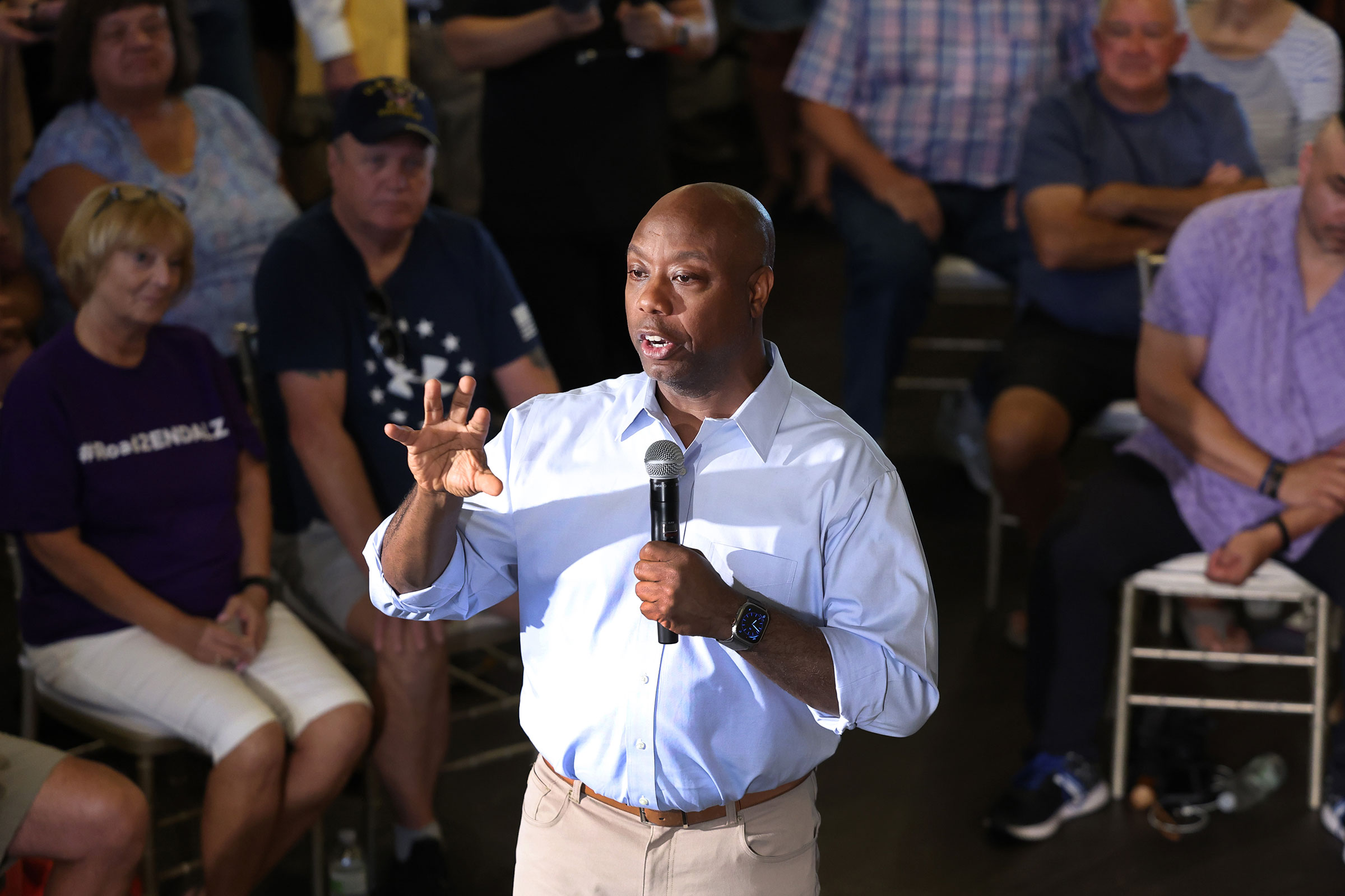 Senator Tim Scott stands in the middle of a group of supporters who are seated as he speaks into a microphone and gestures with his hands