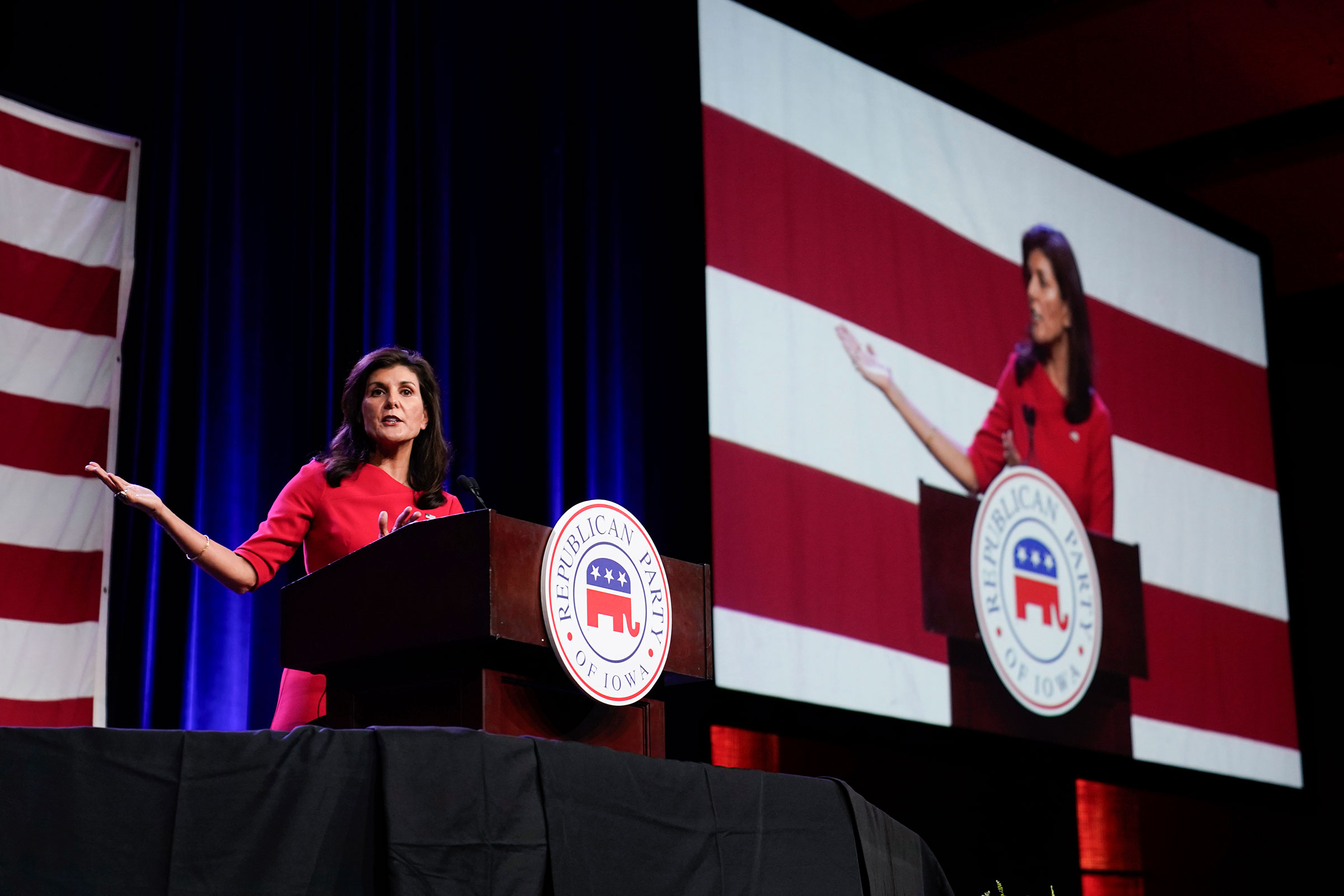 Niki Haley in a red dress gestures and stands at a podium while she speaks with a screen projecting her speaking is seen to her right