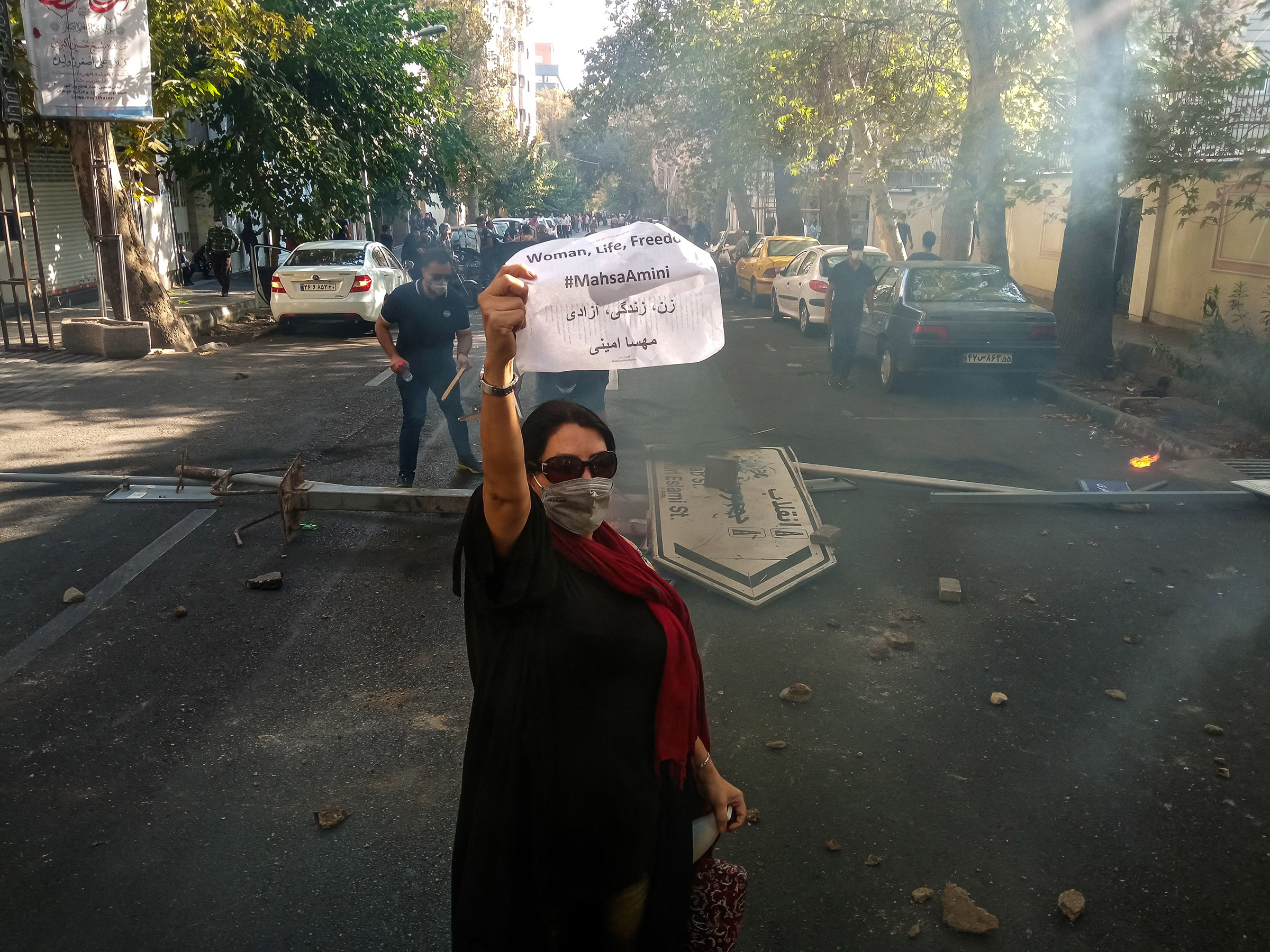 An Iranian woman protester wearing a mask but no hijab holds up a paper reading "Woman, Life, Freedom" and "Mahsa Amini" in front of a street sign on the ground with Revolution and Islamic Republic names on it near Enghelab (Revolution) Street in Tehran, on Oct. 1, 2022.