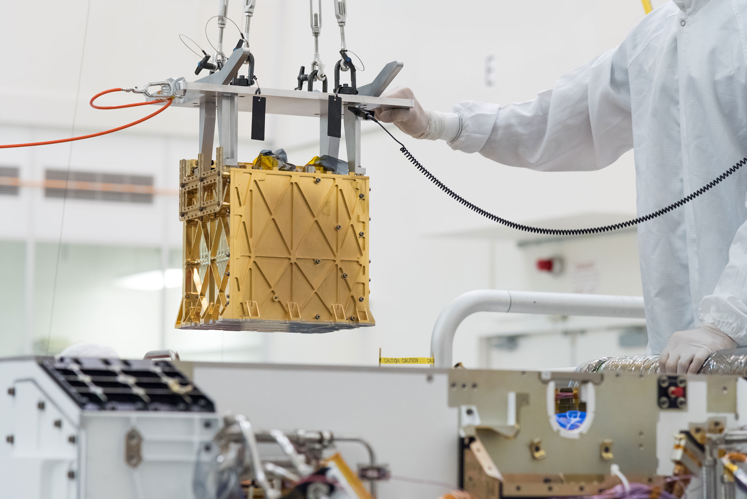 Technicians in the clean room are carefully lowering the Mars Oxygen In-Situ Resource Utilization Experiment (MOXIE) instrument into the belly of the Perseverance rover. (R. Lannom—NASA/JPL-Caltech)