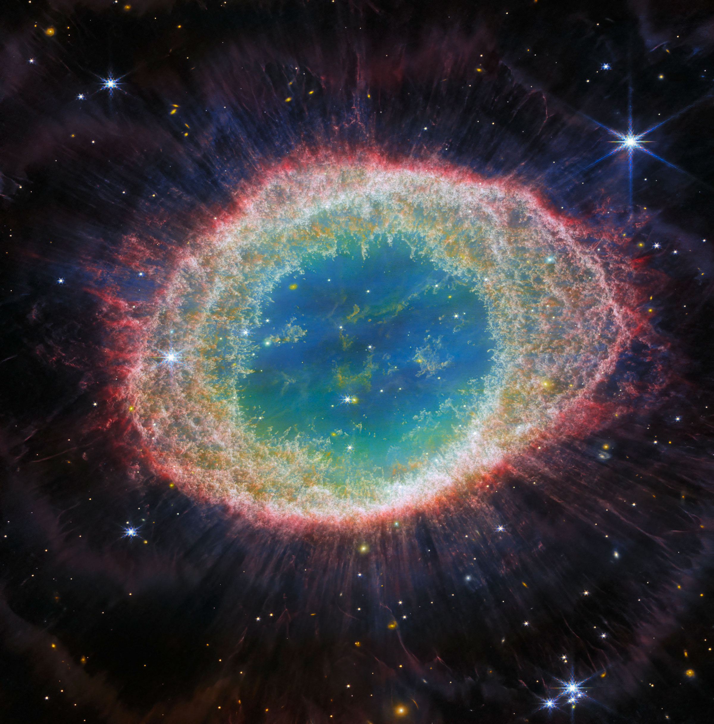 Webb’s near-infrared view of the Ring Nebula has a different color palette. This time, the nebula’s inner cavity hosts shades of blue and green, while the detailed ring transitions through shades of orange in the inner regions and pink in the outer region. Stars litter the scene, with a particularly prominent star with 8 long spikes in the top right corner.
