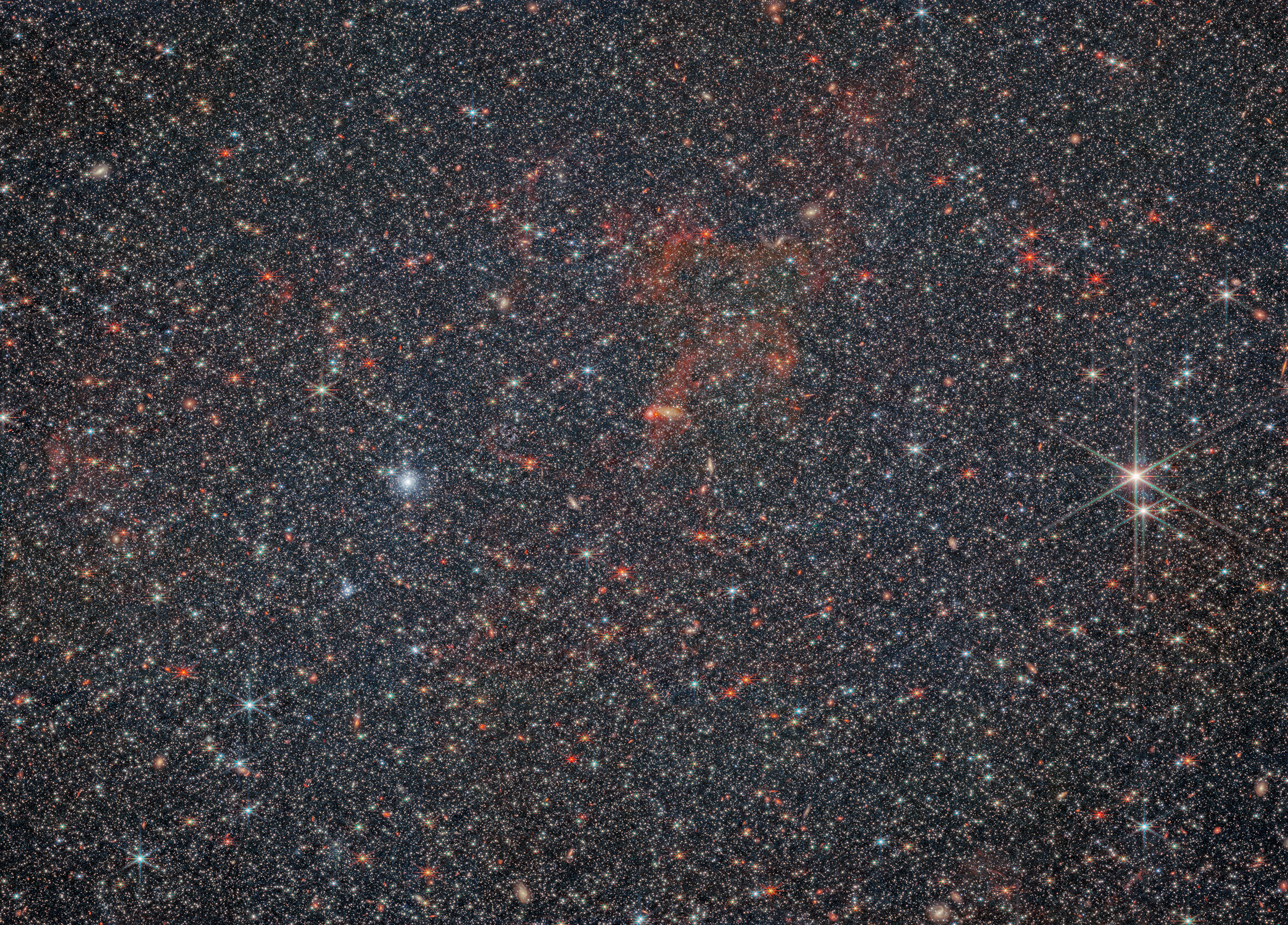 A huge, dense field completely filled with tiny stars. Many galaxies of various shapes and sizes can be seen hiding behind the stars. In the center, there is some faint, wispy, dark red gas. A few of the stars imaged are a bit larger than the rest, with visible diffraction spikes. Two foreground stars are particularly large and bright on the right side.