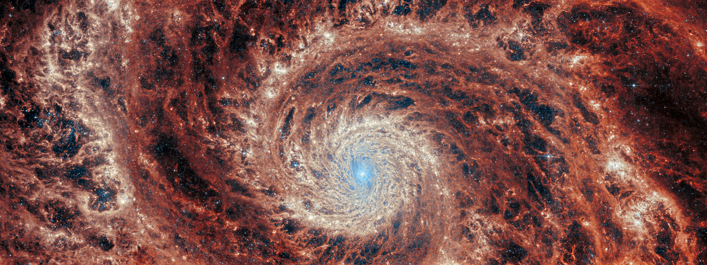 Visão infravermelha média de Webb da galáxia M51.  Uma grande galáxia espiral ocupa toda a imagem.  O núcleo é quase todo branco brilhante, mas também há estruturas detalhadas e rodopiantes que lembram água circulando em um ralo.  Há luz branca e azul clara que emana das estrelas e da poeira no centro do núcleo, mas é fortemente limitada ao núcleo.  Os anéis detalhados apresentam faixas de laranja profundo e cinza turvo, que são intercaladas por regiões vazias mais escuras.