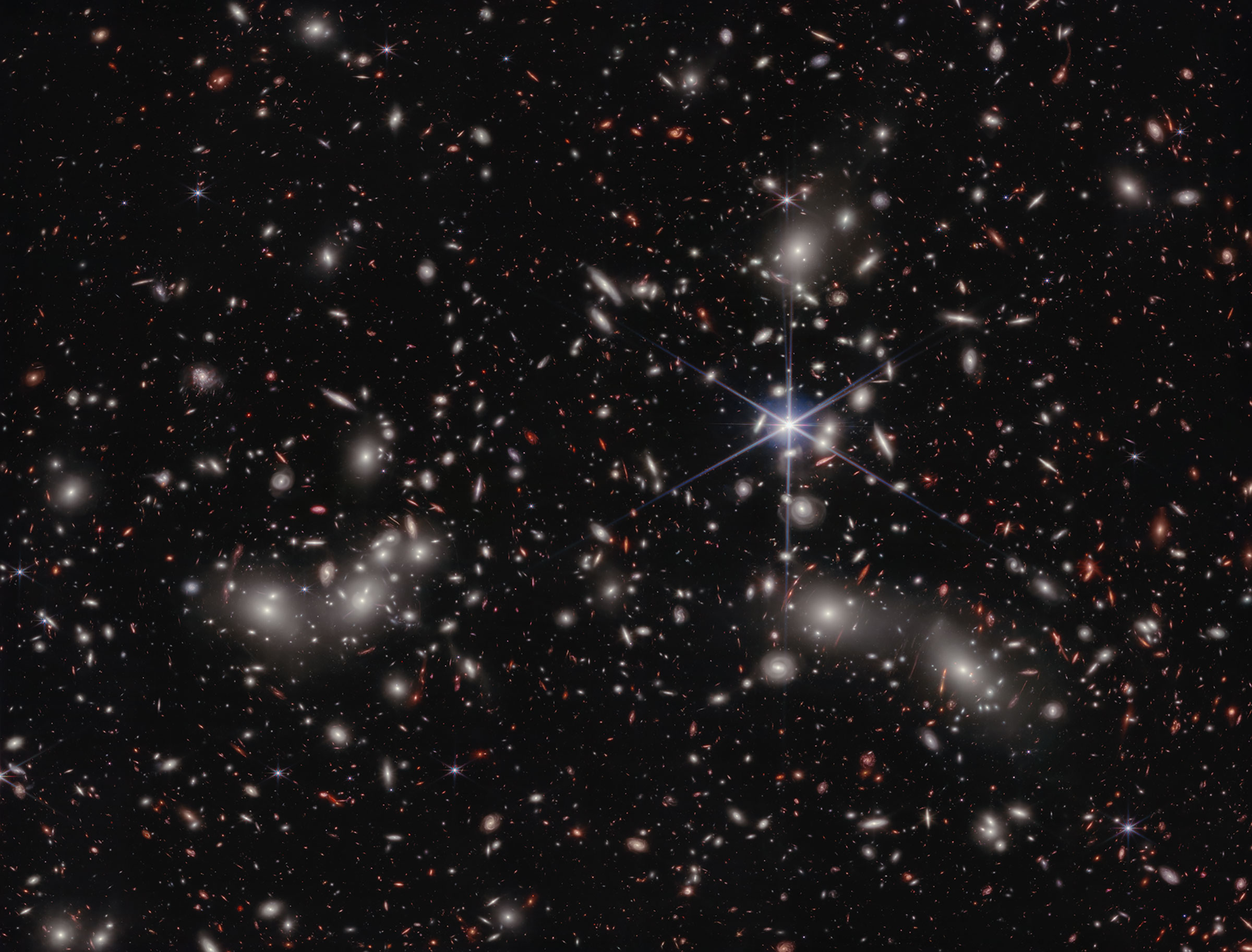 A crowded galaxy field on a black background, with one large star dominating the image just right of center. Three areas are concentrated with larger white hazy blobs on the left, lower right, and upper right above the single star. Scattered between these areas are many smaller sources of light; some also have a hazy white glow, while many other are red or orange. Even without zooming in, different galaxy shapes are detectable, like spirals, ovals, and arcs.