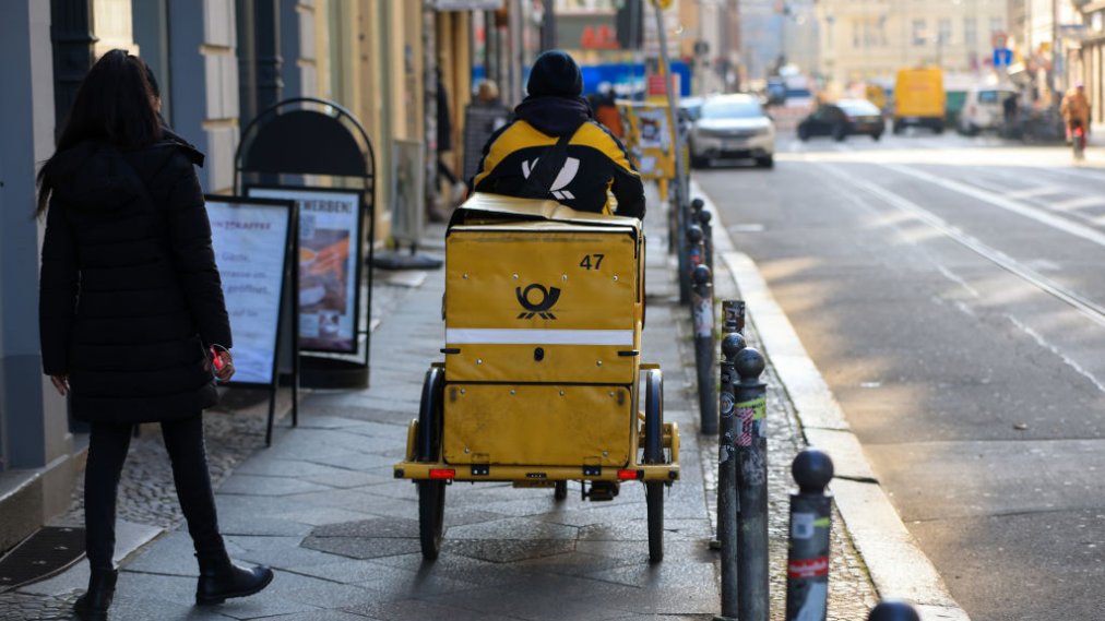 A delivery driver for Deutsche Post AG cycles on a sidewalk.