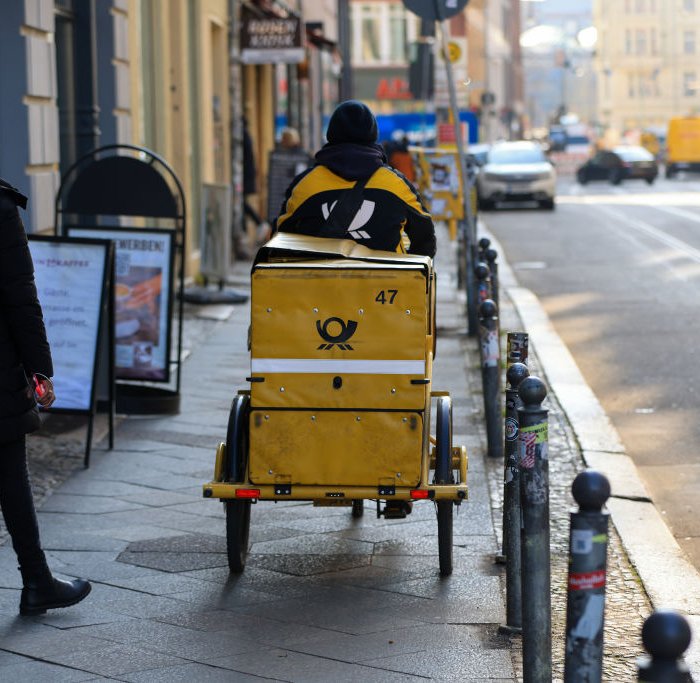 A delivery driver for Deutsche Post AG cycles on a sidewalk.