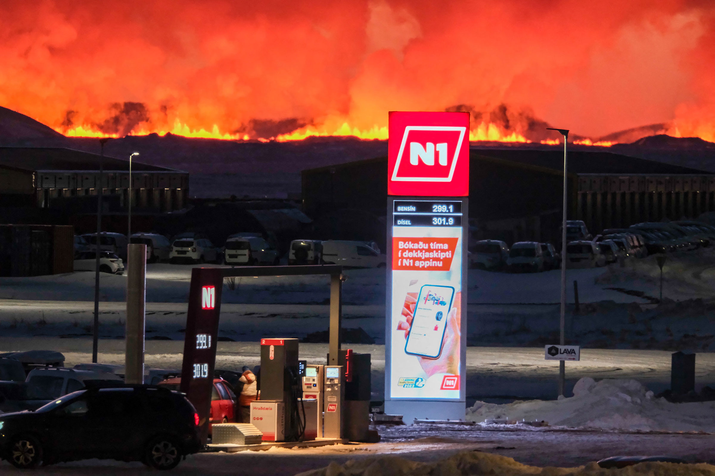 People fill up their vehicles at a petrol station as lava and billowing smoke pours out of a fissure during a volcanic eruption near Grindavík